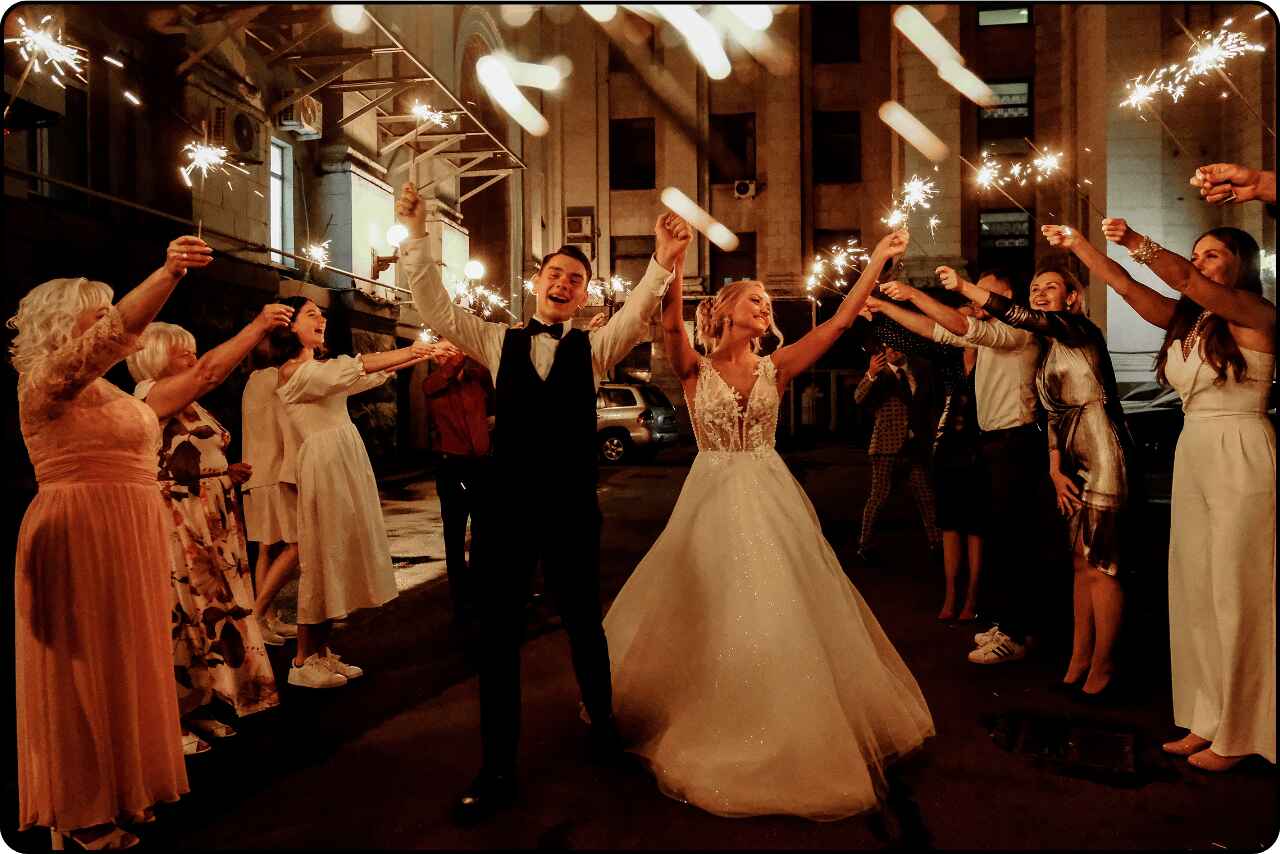 Happy newlywed couple dancing with friends under string lights at a nighttime wedding reception.